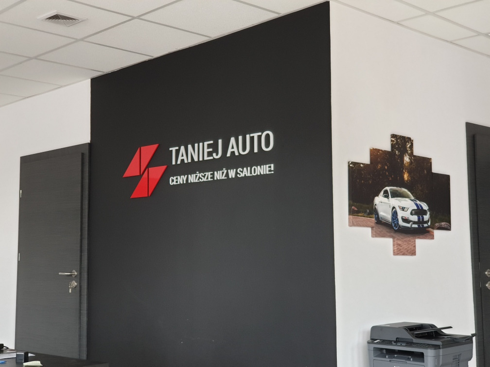 Wnętrze biura firmy 'TANIEJ AUTO' z logo na czarnej ścianie i obrazem sportowego samochodu na ścianie obok, widoczny również fragment drukarki.