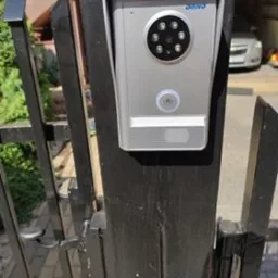 Srebrny wideodomofon zamontowany na czarnym, metalowym słupku bramy wjazdowej. Widoczne diody podczerwieni i przycisk dzwonka. W tle fragment budynku i zaparkowany samochód.