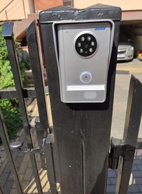 Srebrny wideodomofon zamontowany na czarnym, metalowym słupku bramy wjazdowej. Widoczne diody podczerwieni i przycisk dzwonka. W tle fragment budynku i zaparkowany samochód.