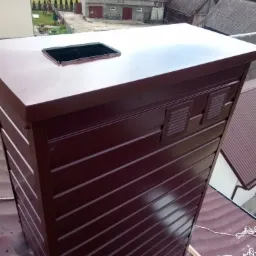 Nowa, brązowa obudowa komina z blachy z otwartym otworem wentylacyjnym na dachu z blachy, widok z góry na tle zabudowań gospodarczych i drzew.