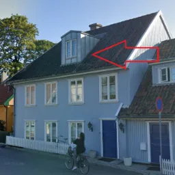 Dwupiętrowy dom z ciemnoczerwonym dachem z dachówki, jasnoniebieską fasadą i lukarną z metalowym obiciem, widziany z ulicy z rowerzystką.