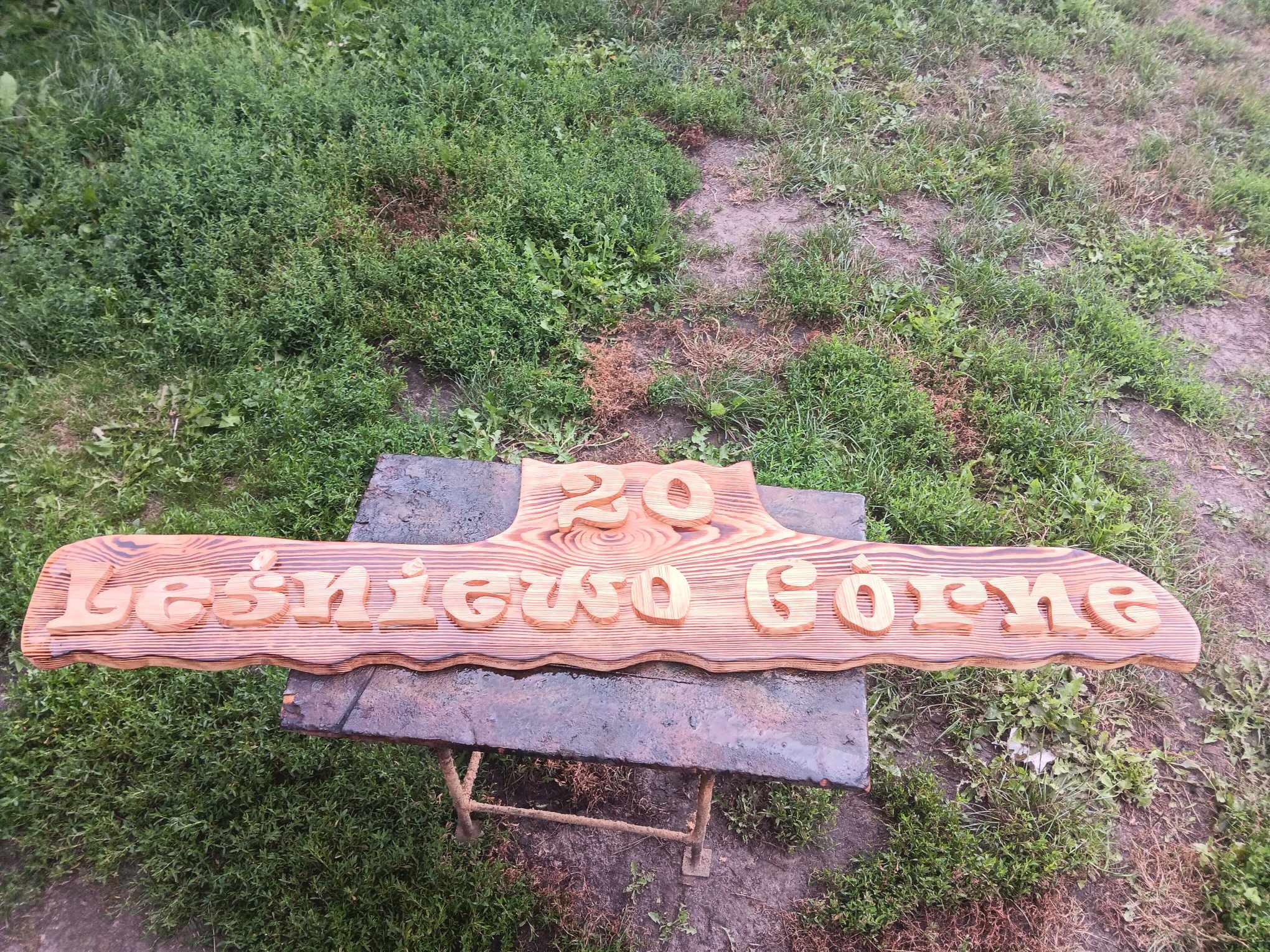 Drewniany szyld z wyciętym napisem 'Leśniewo Górne 20', leżący na metalowym stole w otoczeniu trawy.