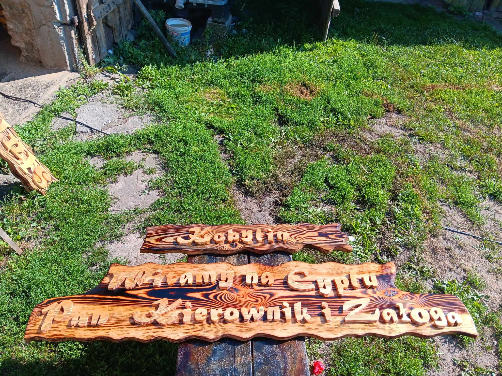 Drewniane tabliczki z wypalonymi napisami 'Kobylin' oraz 'Witamy na Cyplu Pan Kierownik i Załoga' leżą na trawie.