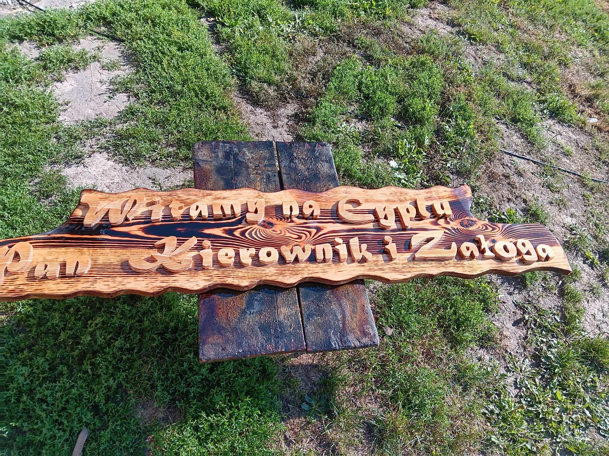 Ręcznie wykonana, drewniana tabliczka z napisem 'Witamy na Cyrku Pan Kierownik i Załoga', umieszczona na dwóch ciemnych, drewnianych klockach na tle trawy.