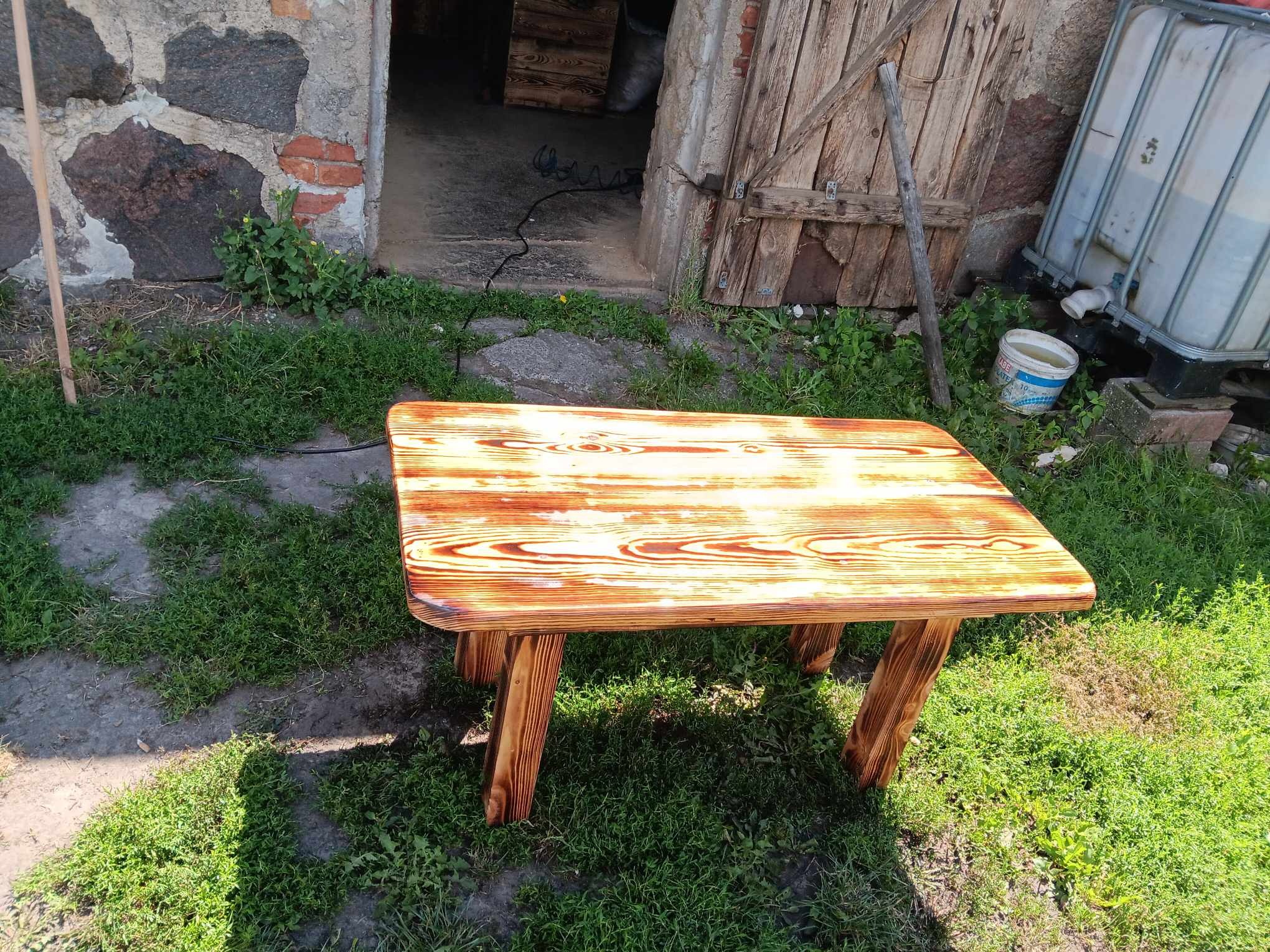 Drewniany stolik z wyraźnym usłojeniem, polakierowany na brązowo-pomarańczowy kolor, stoi na trawie przed budynkiem gospodarczym z kamienia i drewna.