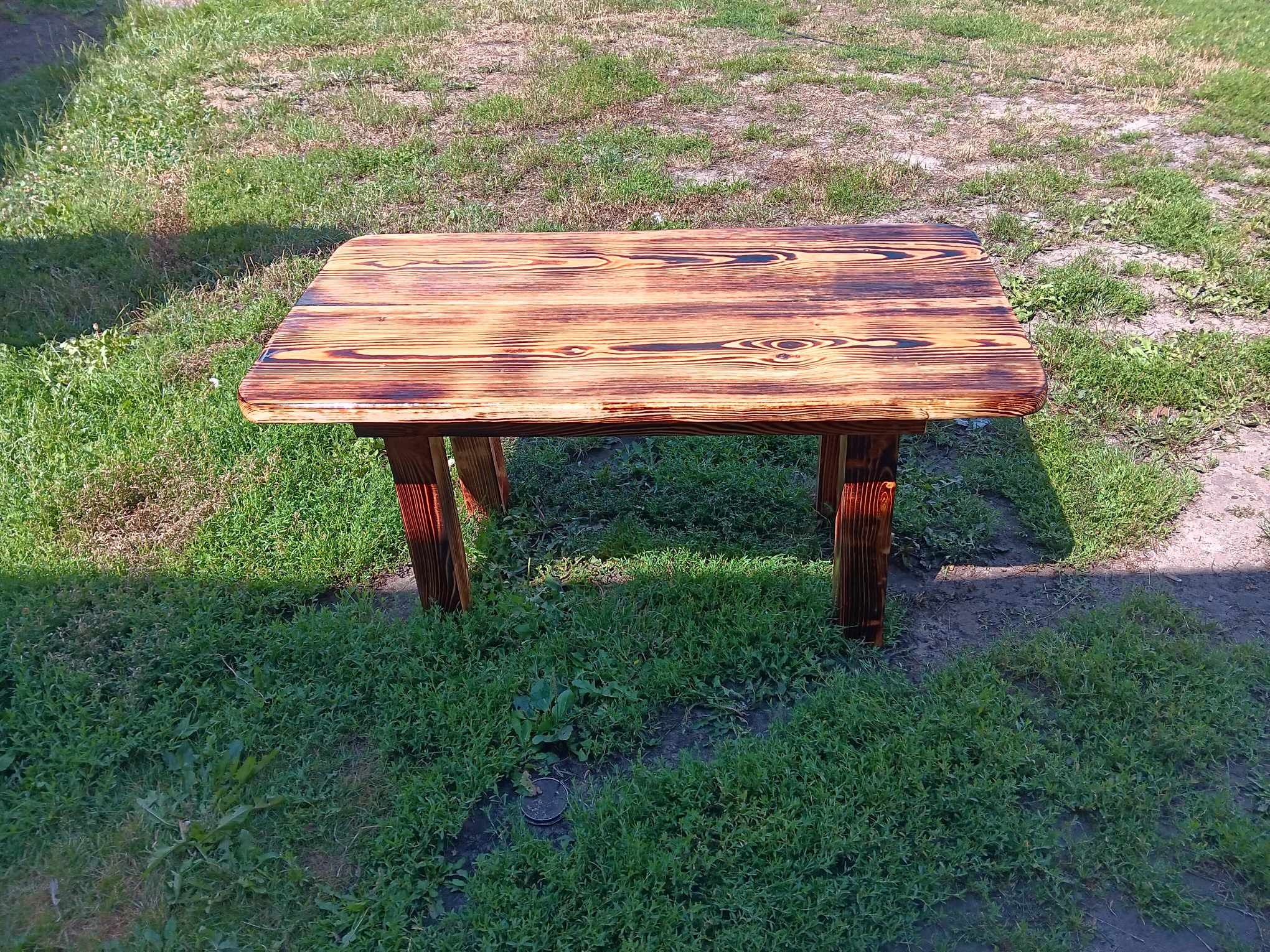 Stolik kawowy z palonej deski, widoczne słoje drewna, ustawiony na trawie.