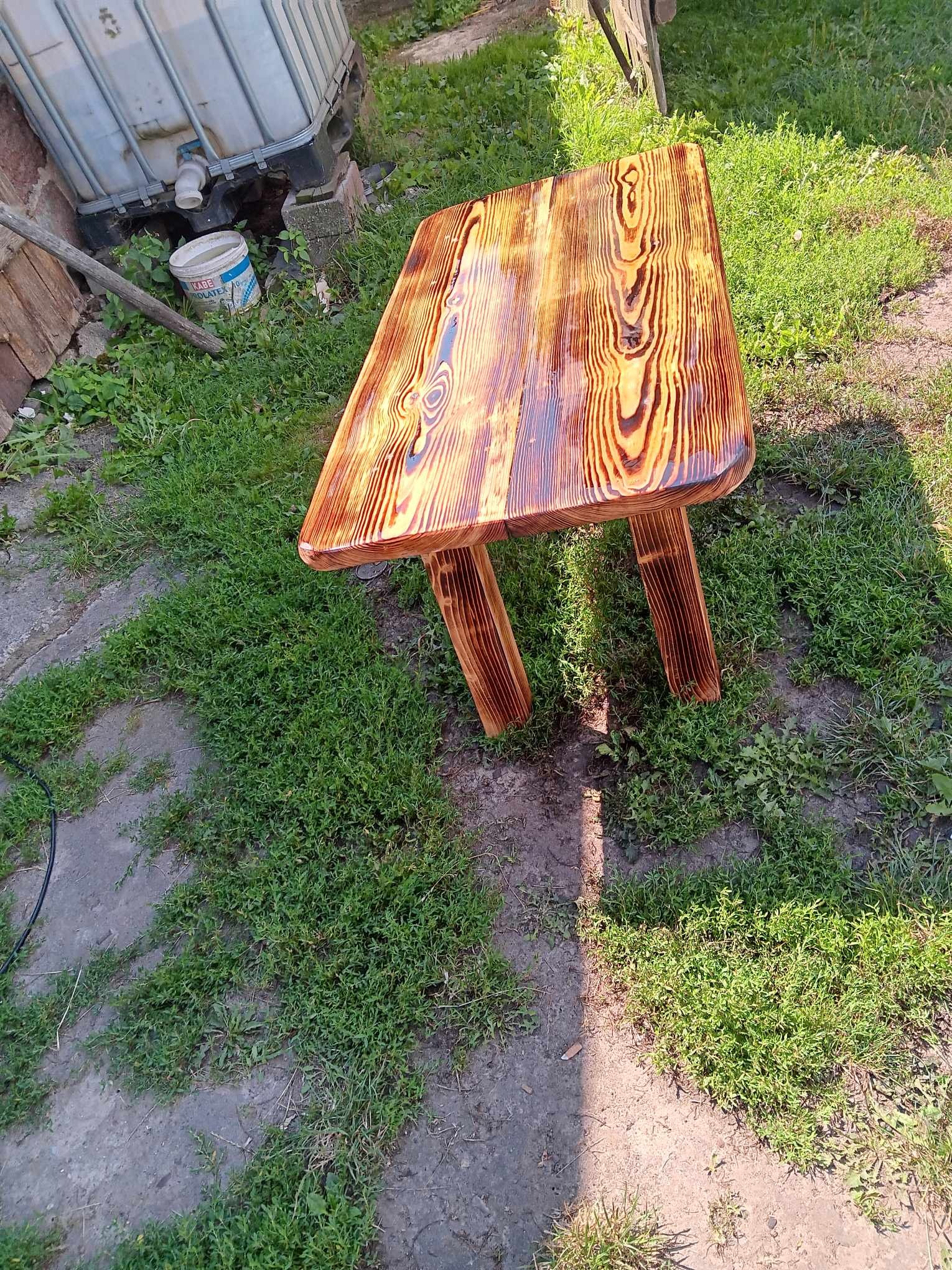 Niewielki, ręcznie wykonany stolik z drewna o wyraźnym usłojeniu, polakierowany na wysoki połysk, ustawiony na trawie w przydomowym ogrodzie, obok betonowej wylewki i plastikowego pojemnika na wodę.