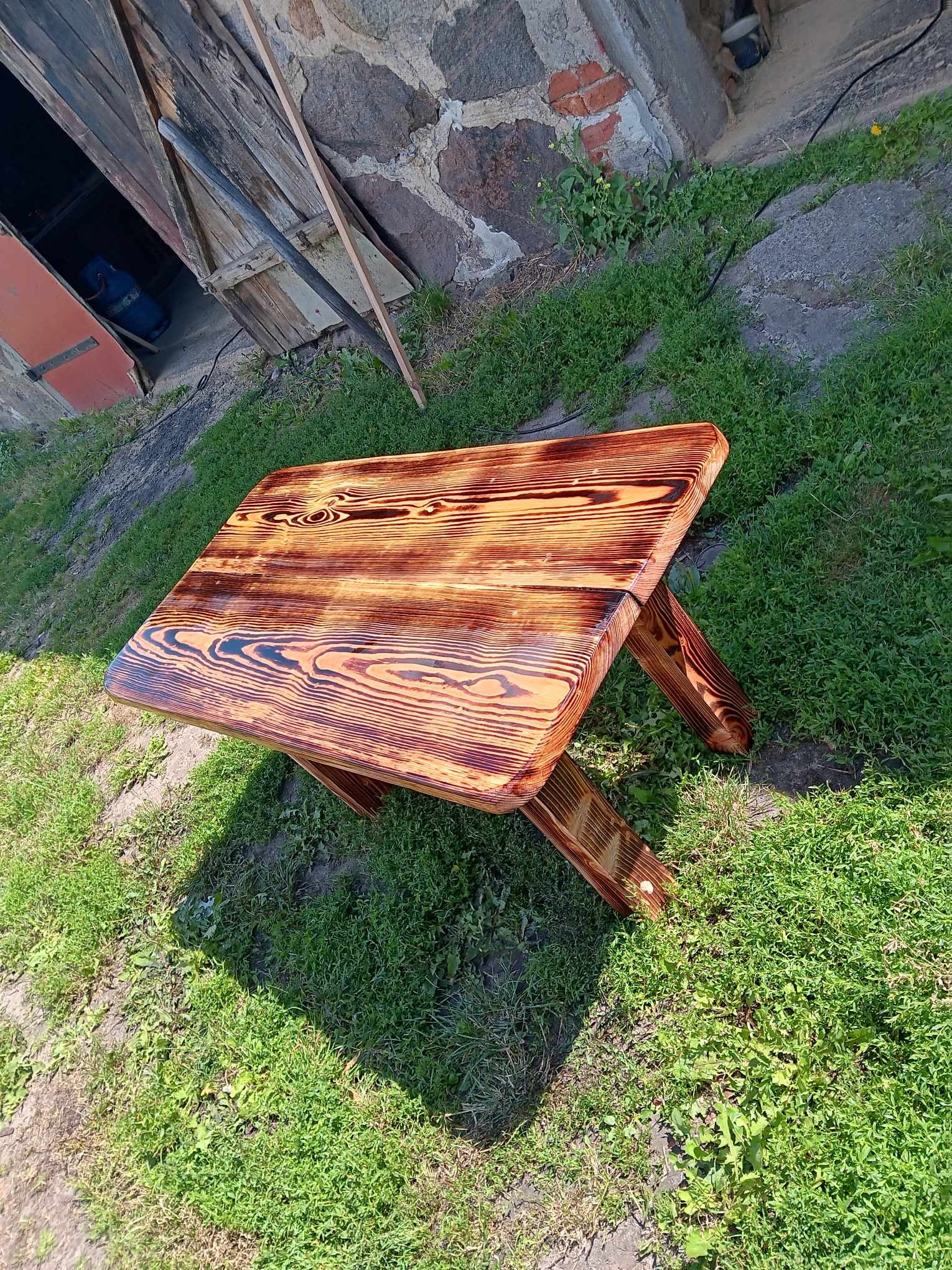 Mały stolik z tarcicy budowlanej, z widocznym usłojeniem drewna, polakierowany na brązowo, stojący na trawie przed budynkiem gospodarczym z kamienia.