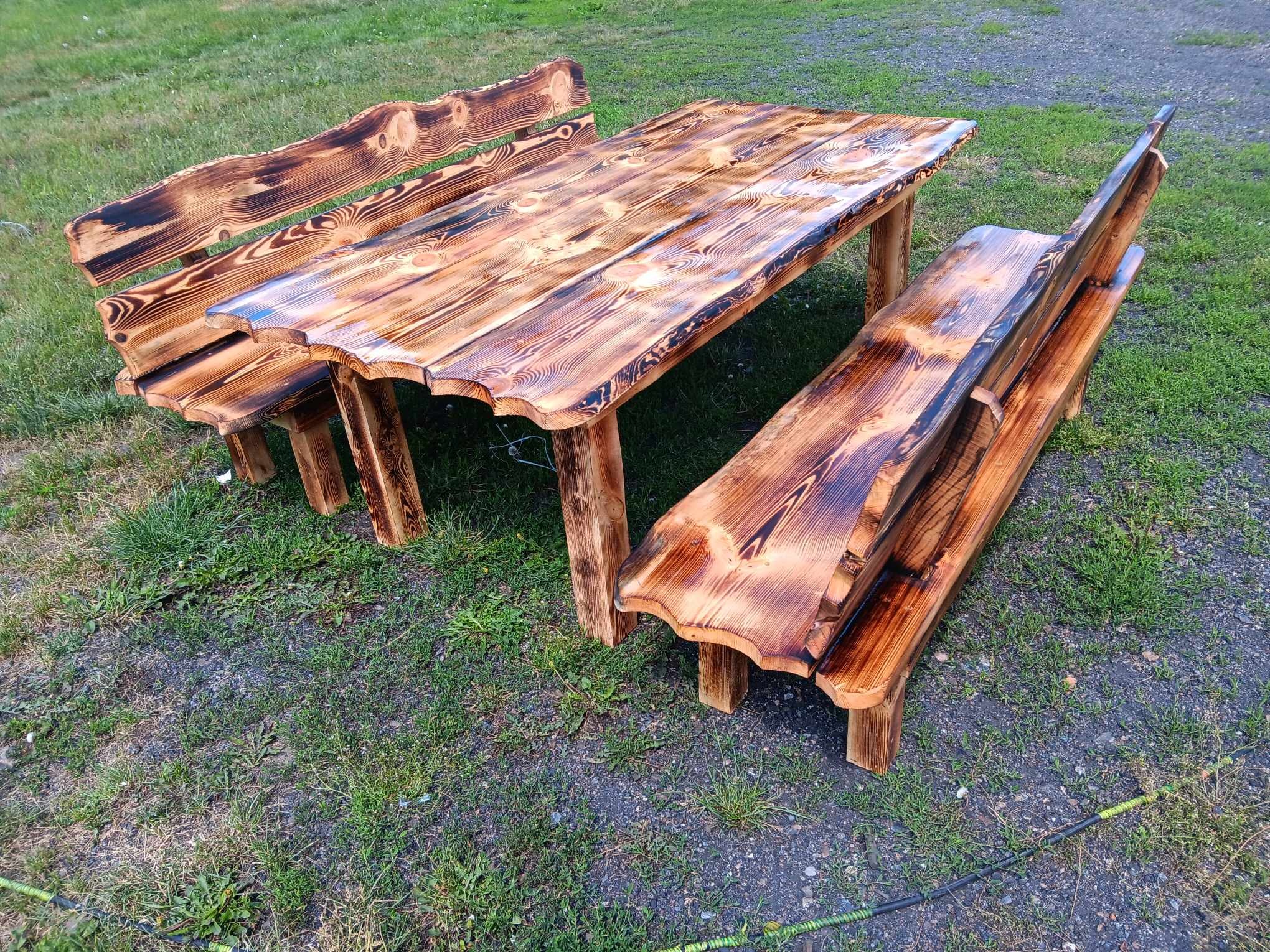 Rustykalny zestaw mebli ogrodowych z drewna, składający się z stołu i dwóch ławek, z wyraźnym rysunkiem słojów i nieregularnymi krawędziami, stojący na trawie z elementami żwiru.