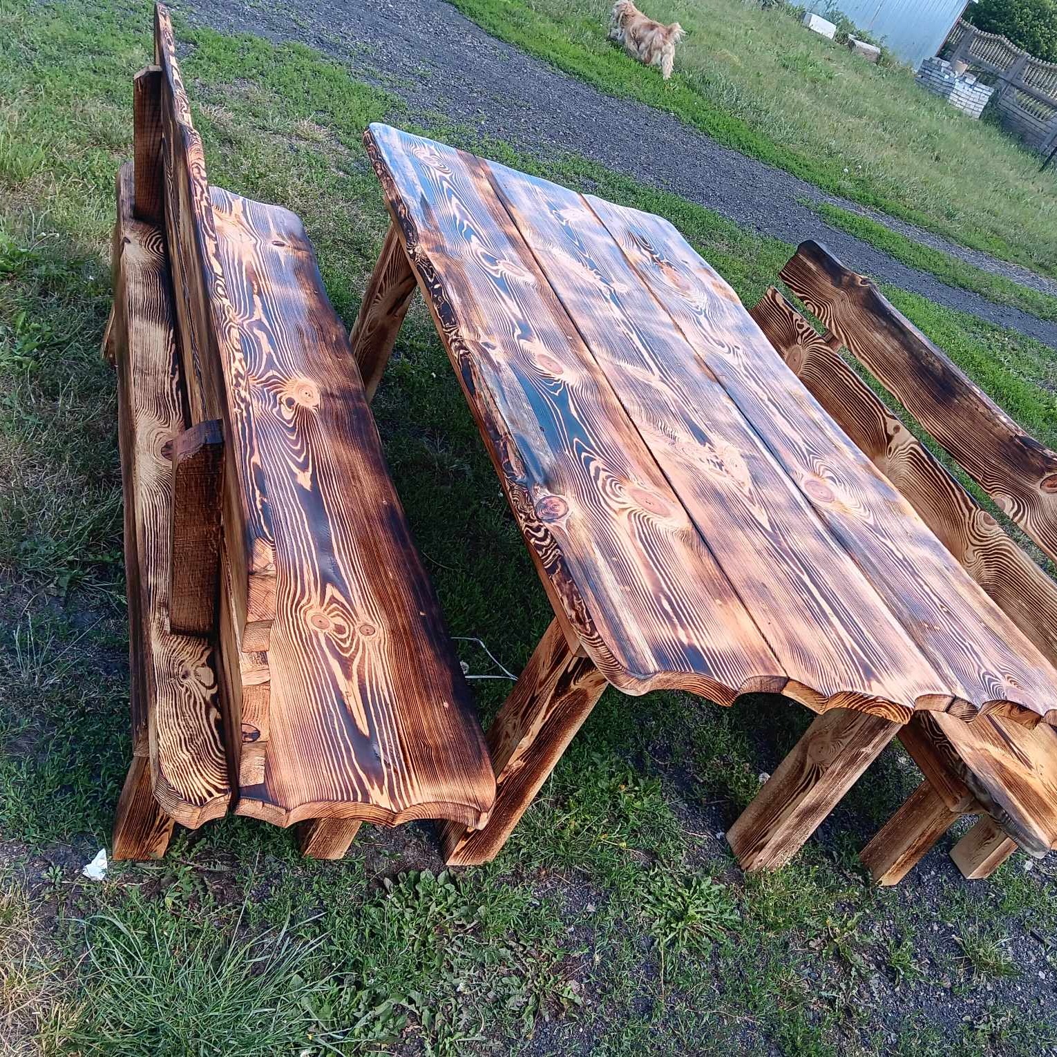 Drewniany stół piknikowy z ławkami, opalany i lakierowany, ustawiony na trawie, w tle pies i budynek gospodarczy.