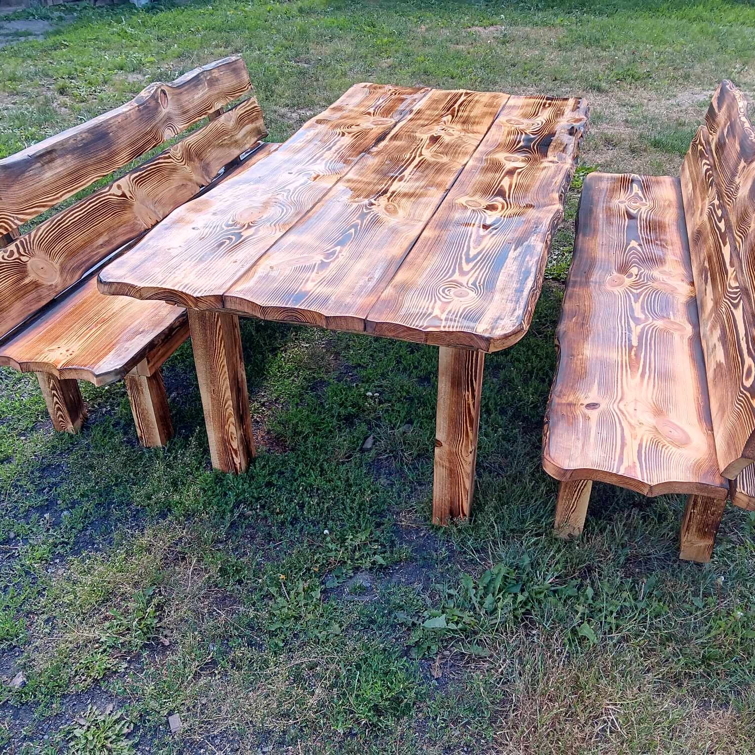 Rustykalny zestaw mebli ogrodowych z surowego drewna: stół i dwie ławki, z wyraźnym usłojeniem i naturalnymi krawędziami, ustawiony na trawie.