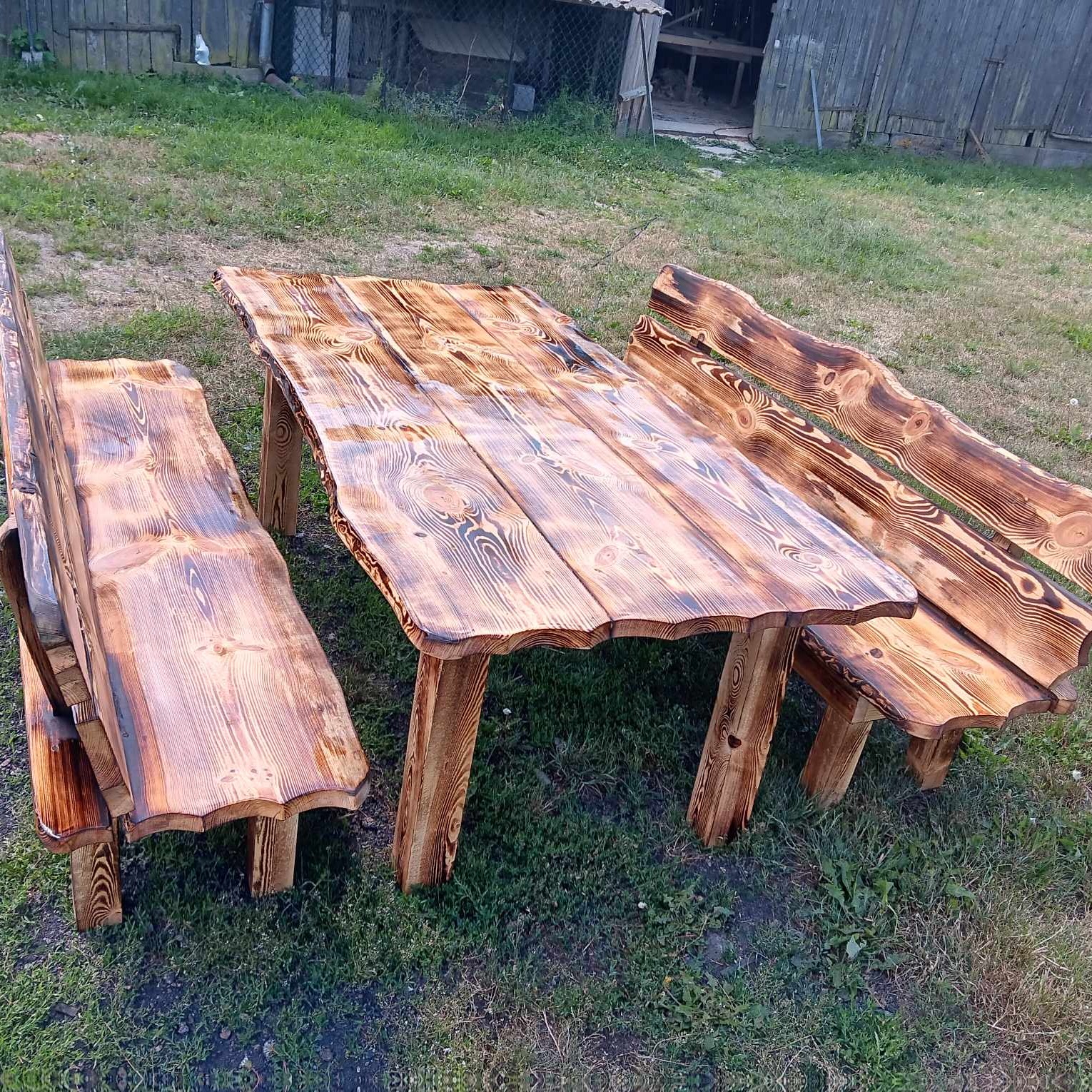 Drewniany stół i dwie ławki z opalonymi brzegami, ustawione na trawie przed drewnianym budynkiem gospodarczym.