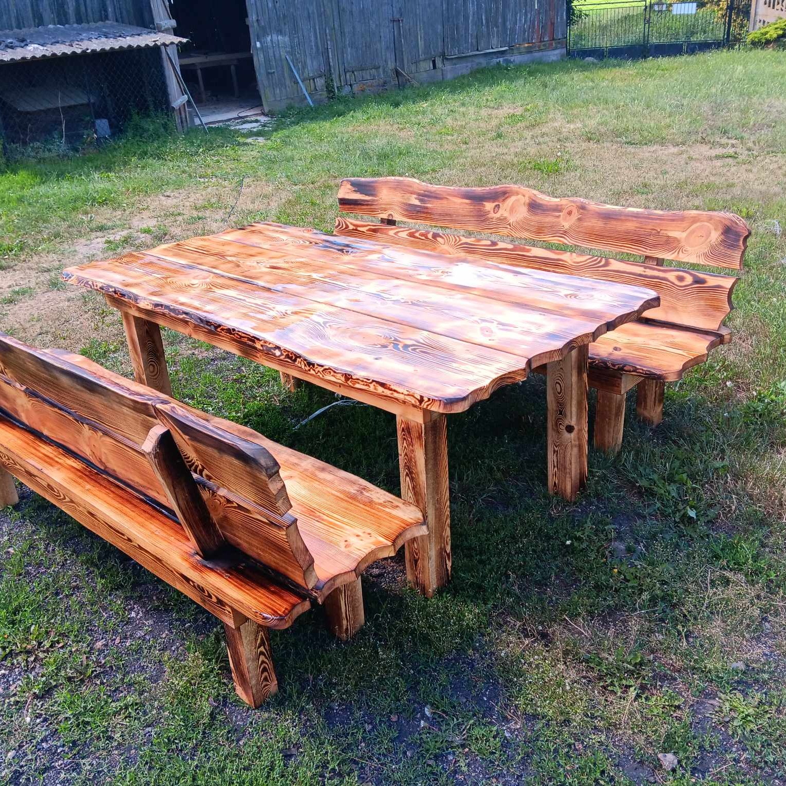 Rustykalny zestaw mebli ogrodowych: stół z blatem o nieregularnym kształcie i dwie ławki z oparciami, wykonane z opalizowanego drewna na tle zielonej trawy i drewnianej stodoły.