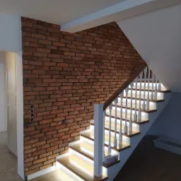 Murowana ściana z cegły klinkierowej sąsiadująca z drewnianymi schodami z białymi balustradami i podświetleniem LED pod stopniami.