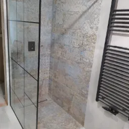 Nowoczesna kabina prysznicowa z czarnymi profilami, zdobione płytki ceramiczne w odcieniach beżu i błękitu, grzejnik drabinkowy w łazience.