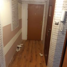 Stiver-Bud - Korytarz w trakcie remontu: pomalowane ściany w pasy, częściowo wykończone ściany z cegły, dwie brązowe drzwi, śmieci na podłodze.
