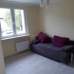 Stiver-Bud - Jasne wnętrze pokoju z szarą sofą i bordowymi poduszkami, widok na okno z grzejnikiem pod nim, oraz nowoczesną lampą sufitową.