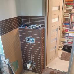 Stiver-Bud - Niedokończona łazienka w trakcie remontu: częściowo położone brązowe płytki, odsłonięte rury, narzędzia na zabudowie stelaża podtynkowego WC, gruz i resztki materiałów w wiadrze, oraz widoczna...