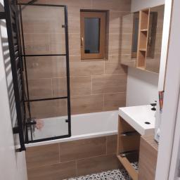 Stiver-Bud - Ukończona łazienka z wanną za szklaną ścianką, umywalką z szafką i lustrem, wykończona płytkami imitującymi drewno i wzorzystą podłogą.