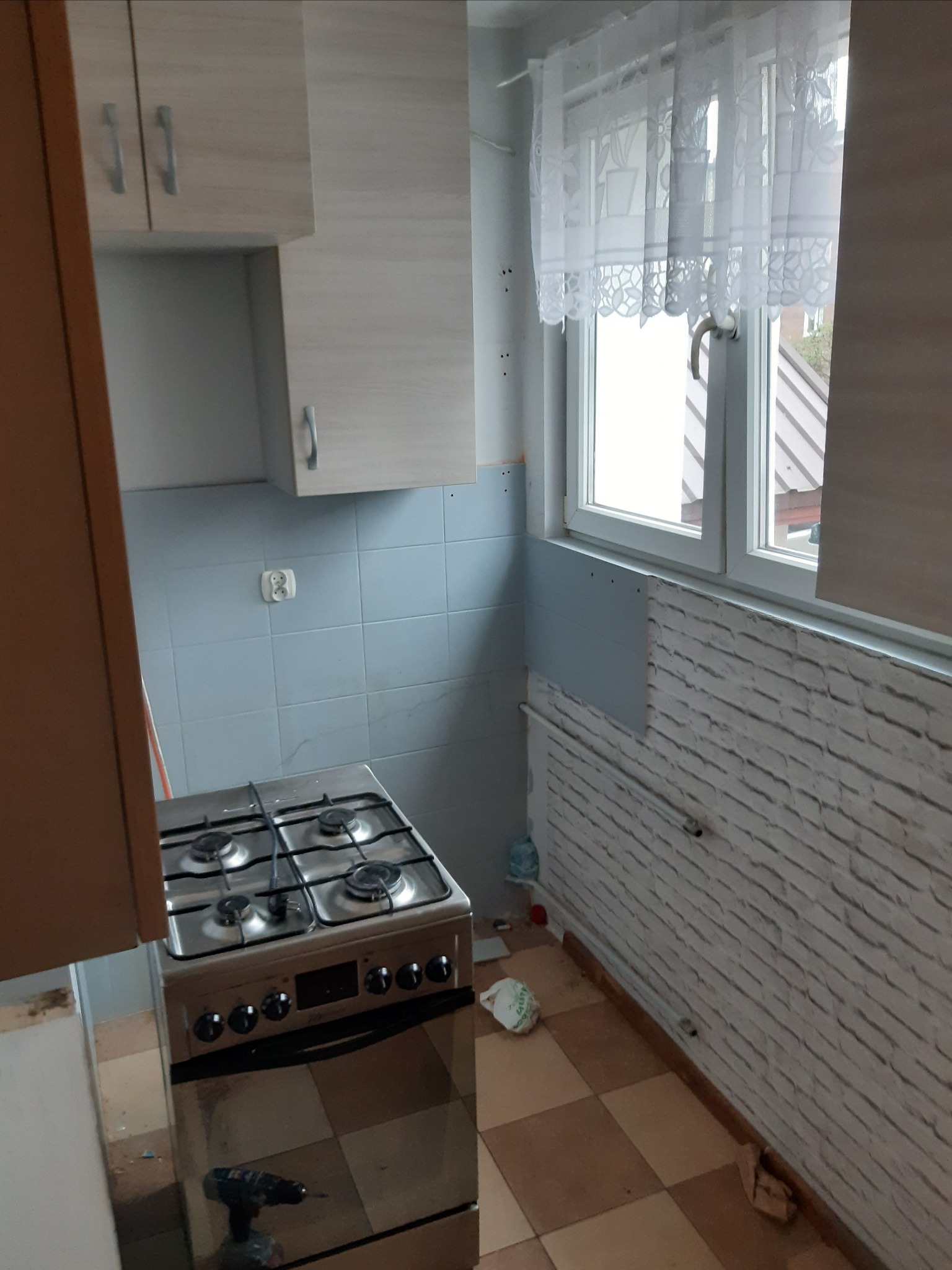 Fragment kuchni w trakcie remontu: widoczna kuchenka gazowa, szafki, okno z firanką, częściowo wykończona ściana z imitacją cegły i narzędzie - wiertarka.