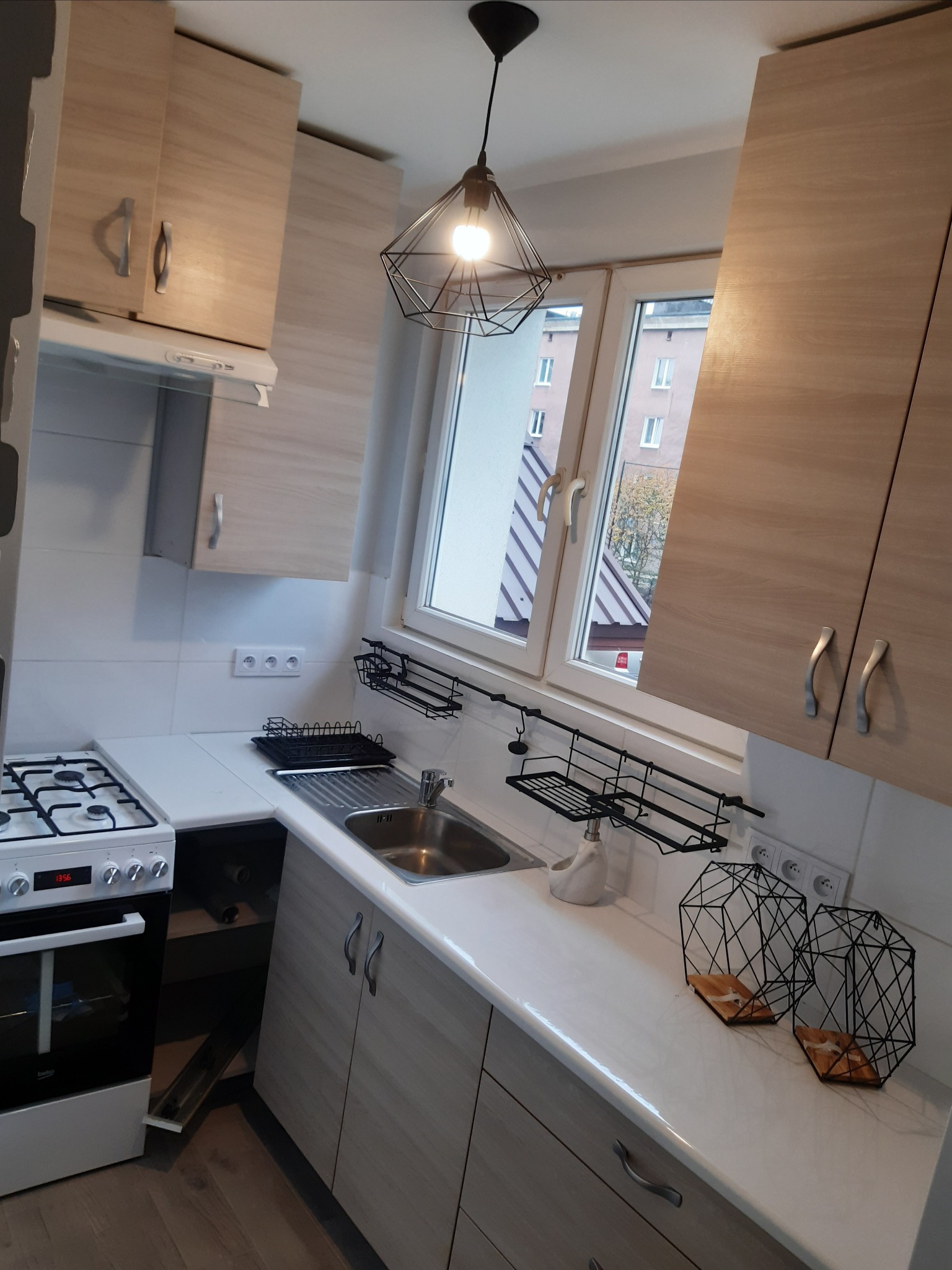 Jasna kuchnia z drewnianymi frontami, białym blatem i geometryczną lampą wiszącą, widok na okno zza zlewu.