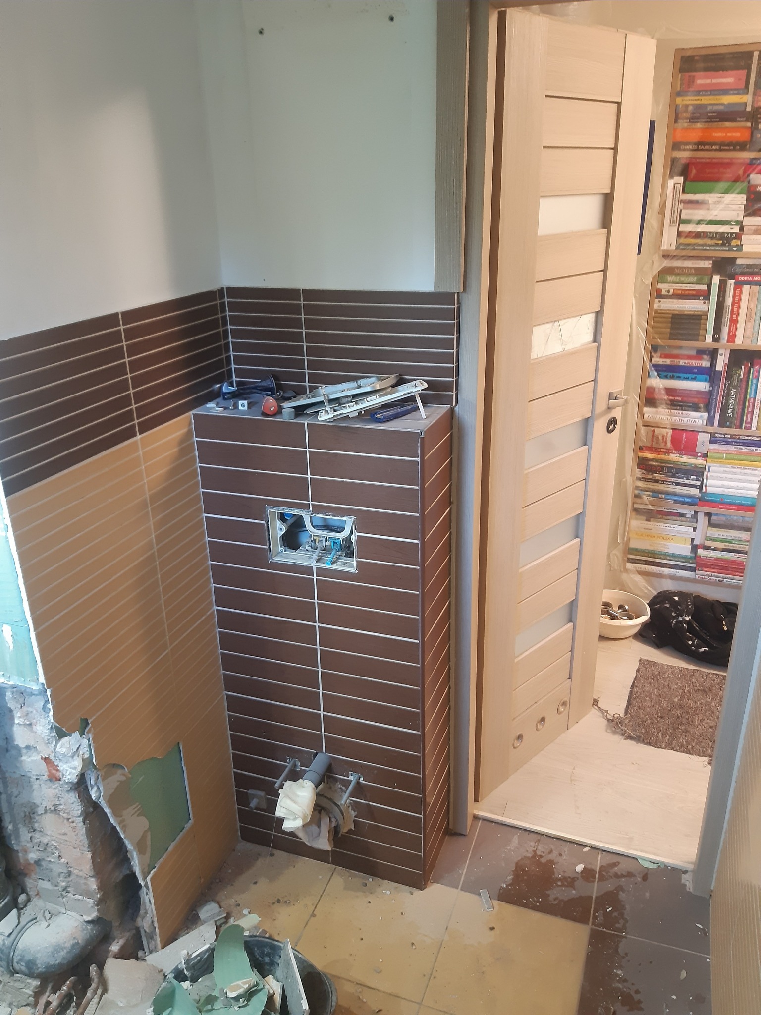 Niedokończona łazienka w trakcie remontu: częściowo położone brązowe płytki, odsłonięte rury, narzędzia na zabudowie stelaża podtynkowego WC, gruz i resztki materiałów w wiadrze, oraz widoczna...