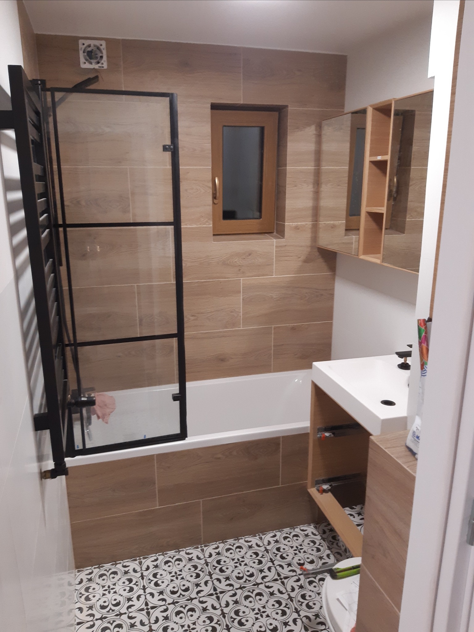 Ukończona łazienka z wanną za szklaną ścianką, umywalką z szafką i lustrem, wykończona płytkami imitującymi drewno i wzorzystą podłogą.