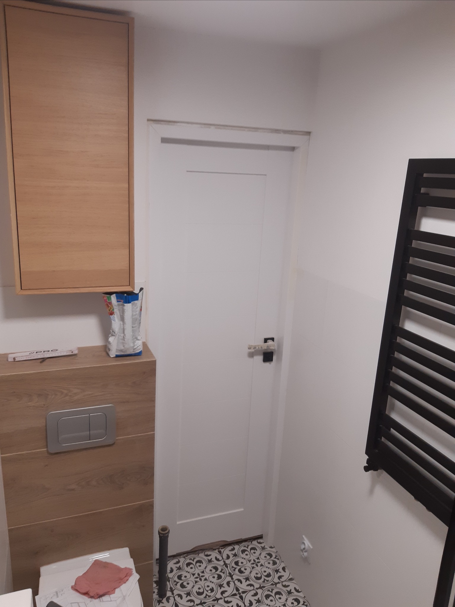 Nowoczesna łazienka po remoncie: wisząca szafka z drewnopodobnym frontem, toaleta w zabudowie, czarno-biała mozaika na podłodze, białe drzwi i grzejnik drabinkowy w kolorze czarnym.