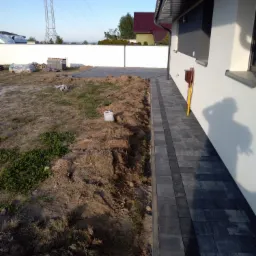 Fragment posesji w trakcie prac brukarskich: ułożona ścieżka z ciemnej kostki obok ściany budynku, w tle palety z kostką, wykopana ziemia i słup wysokiego napięcia.