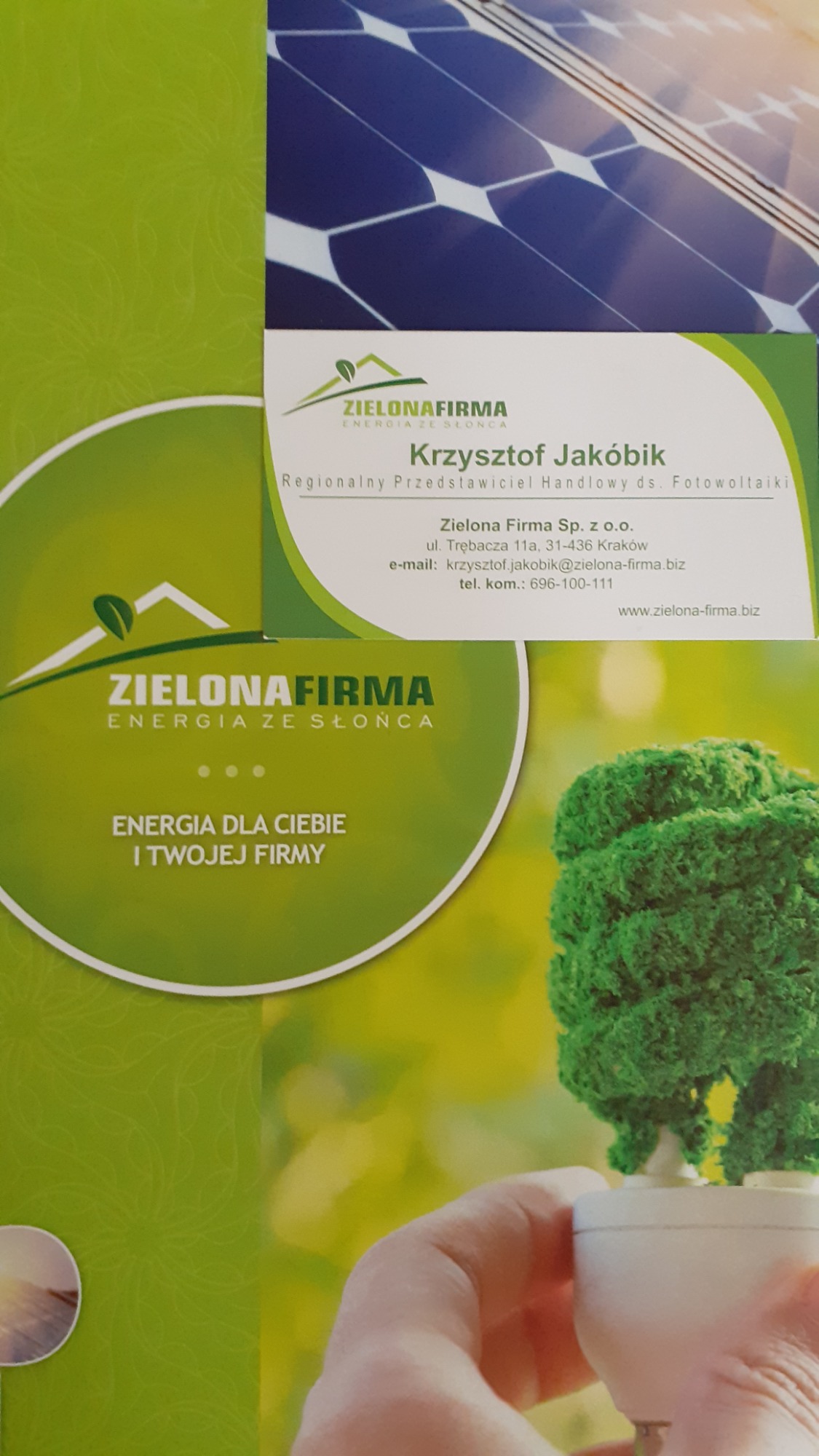 Ulotka reklamowa firmy Zielona Firma z wizytówką Krzysztofa Jakóbika, przedstawiciela handlowego ds. fotowoltaiki, na tle paneli słonecznych i żarówki z zielonym krzewem.