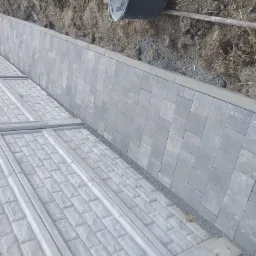 Świeżo ułożona szara kostka brukowa tworząca wąski chodnik, oddzielona od ziemi obrzeżem, z widocznymi elementami konstrukcyjnymi po lewej stronie.