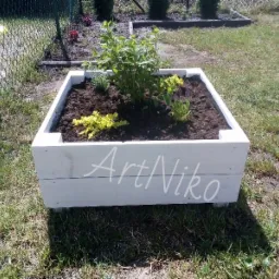 Biała, drewniana skrzynia na rośliny z napisem 'ArtNiko', wypełniona ziemią i sadzonkami, ustawiona na trawniku w ogrodzie.