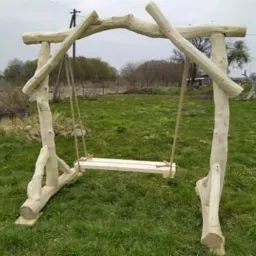 Drewniana huśtawka ogrodowa wykonana z naturalnych gałęzi, umieszczona na trawniku, widoczny słup energetyczny w tle.