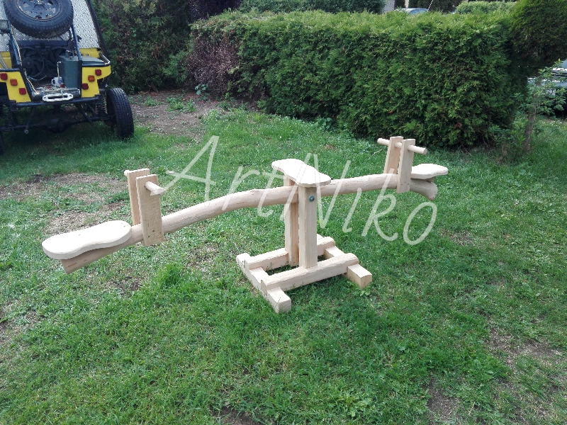 Drewniana huśtawka wahadłowa dla dzieci, wykonana z jasnego drewna, ustawiona na trawniku w ogrodzie, z żółtym pojazdem terenowym w tle.