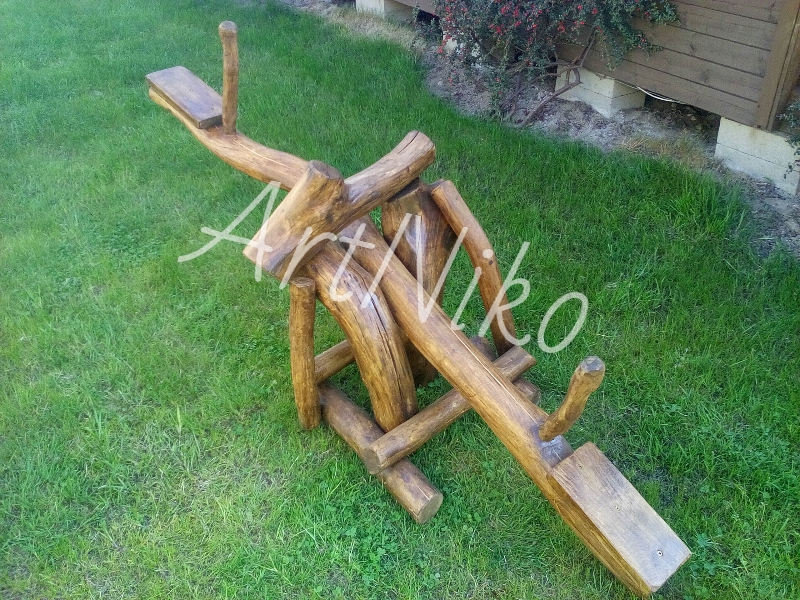 Drewniana huśtawka wagowa w stylu rustykalnym, wykonana z okrąglaków, ustawiona na trawniku przed drewnianym budynkiem.