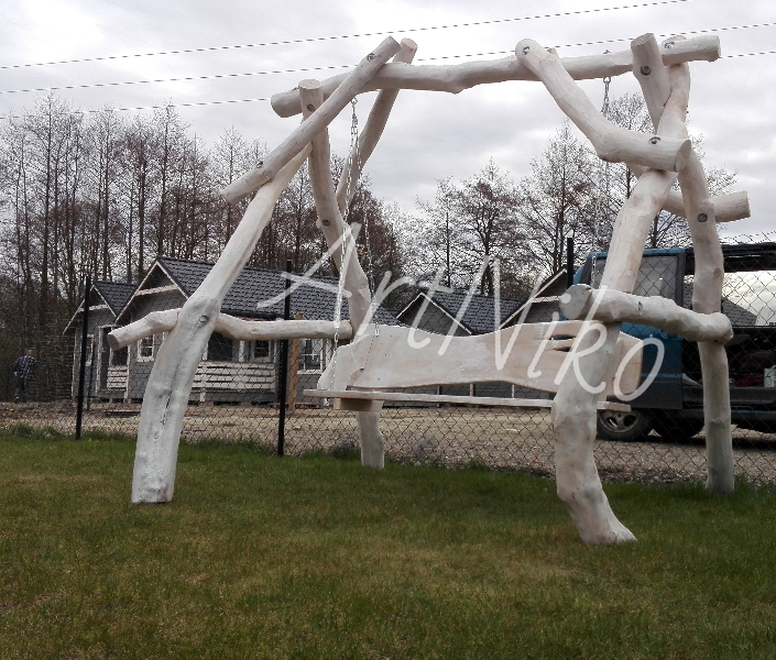 Drewniana huśtawka ogrodowa w stylu rustykalnym, wykonana z pomalowanych na biało gałęzi, na tle zielonego trawnika, z widocznymi domkami letniskowymi w oddali i zaparkowanym samochodem.