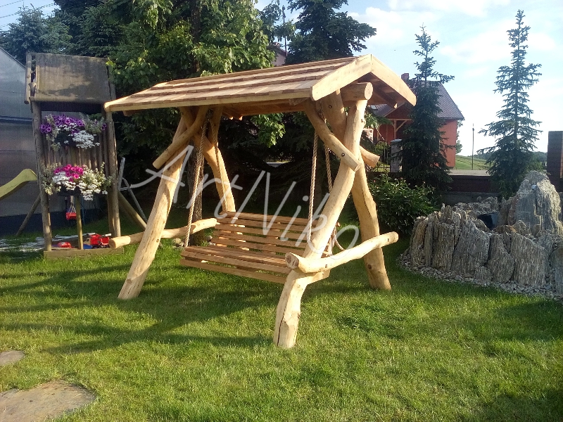 Drewniana huśtawka ogrodowa z zadaszeniem, wykonana z surowych pni, na tle zielonego trawnika i drzew iglastych.