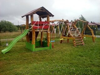 Drewniany plac zabaw z zadaszoną wieżą, zjeżdżalnią, huśtawkami i drabinką, otoczony trawnikiem. Widoczne elementy pomalowane na zielono i czerwono.