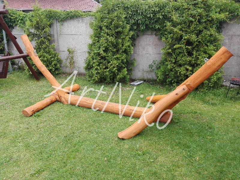 Drewniana konstrukcja huśtawki, wykonana z surowych bali, rozłożona na trawie przed betonowym ogrodzeniem z roślinnością.