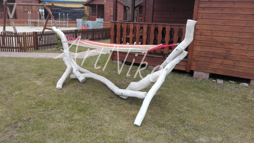 Ręcznie robiony, biały, drewniany stelaż do hamaka z kolorowym hamakiem, ustawiony na trawniku przed drewnianym domkiem.