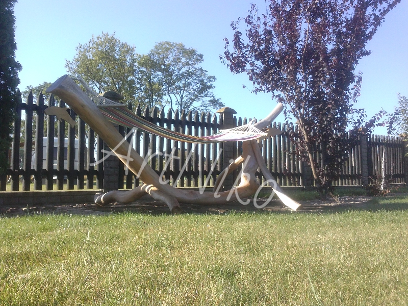 Hamak zawieszony na konstrukcji z naturalnego drewna, ustawiony na trawniku przed drewnianym ogrodzeniem i drzewami.