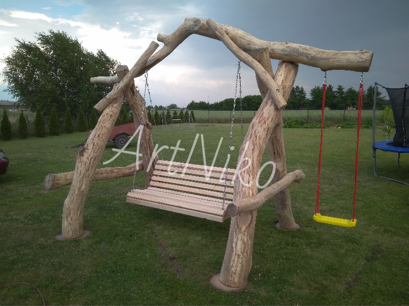 Drewniana huśtawka ogrodowa z bali i desek, zawieszona na łańcuchach i czerwonych linach, z żółtym siedziskiem, na tle zielonego trawnika i pochmurnego nieba.