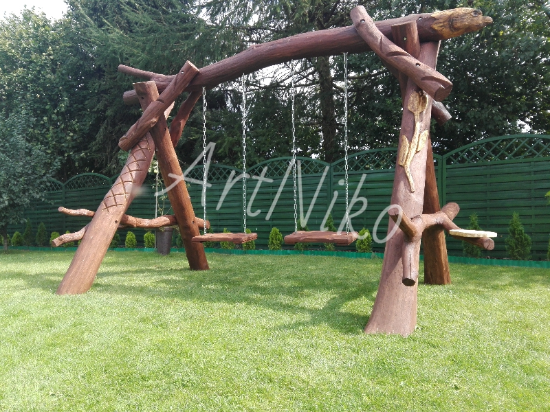 Drewniana huśtawka ogrodowa z rzeźbionymi elementami, stylizowana na naturalne gałęzie i pnie drzew, z siedziskami zawieszonymi na łańcuchach, na tle zielonego trawnika i ogrodzenia.