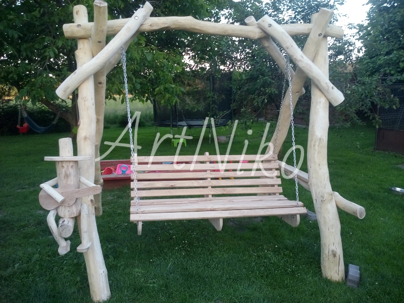 Drewniana huśtawka ogrodowa z rzeźbą grającego ludzika, zawieszona na łańcuchach, na tle trawnika i drzew