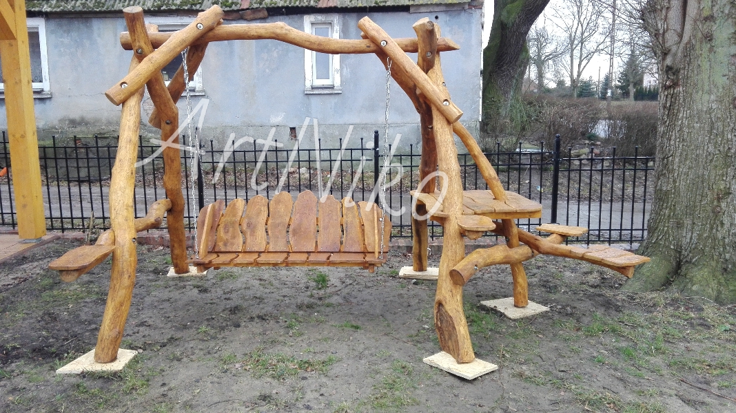 Drewniana huśtawka ogrodowa z elementami siedzisk i stolików, wykonana z naturalnych gałęzi i pni, osadzona na betonowych płytach w ogrodzie.