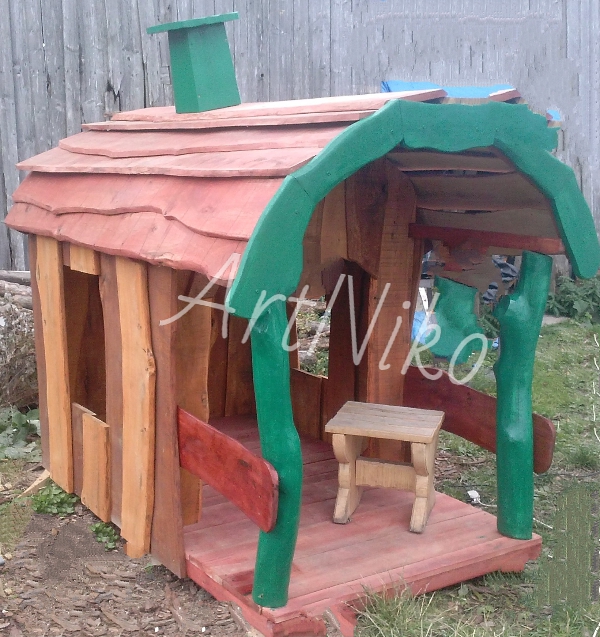 Mała drewniana altana dla dzieci z czerwonym dachem i zielonymi elementami dekoracyjnymi, z małym stołkiem w środku, na tle drewnianego płotu.