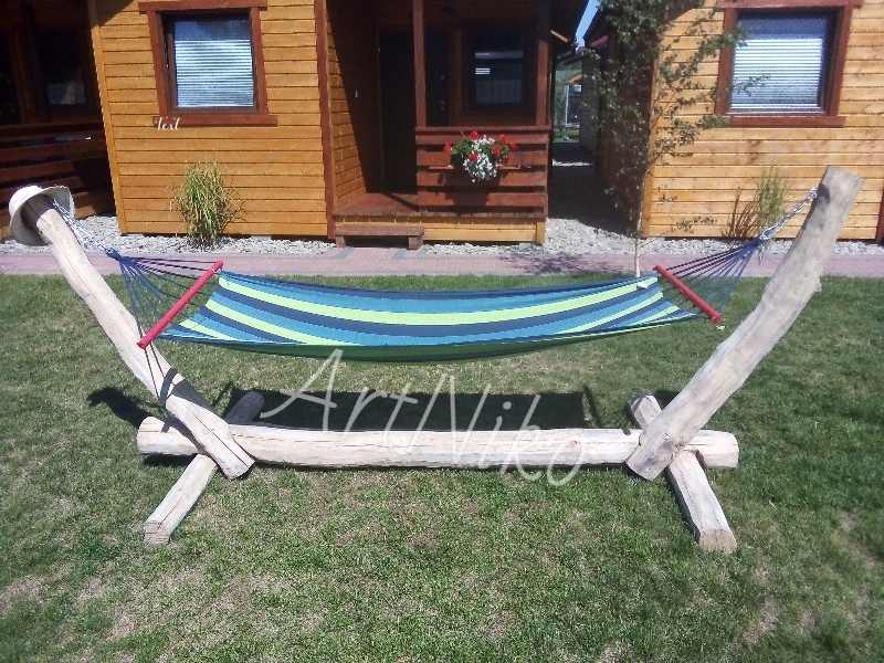 Ręcznie robiony hamak z drewna, z niebiesko-zielonym pasiakowanym materiałem do leżenia, ustawiony na trawie przed drewnianymi domkami letniskowymi.