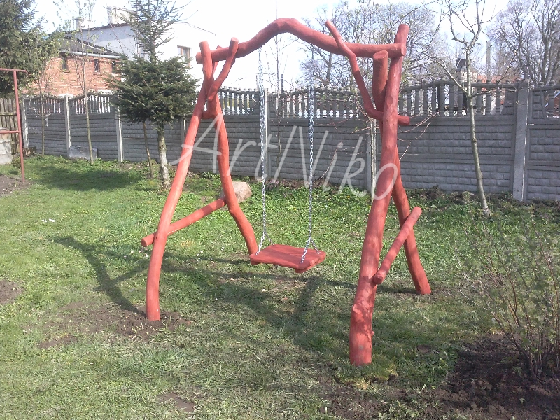 Ręcznie robiona huśtawka ogrodowa z drewna pomalowanego na czerwono, zawieszona na metalowych łańcuchach, tło stanowi zielony trawnik i betonowy płot.