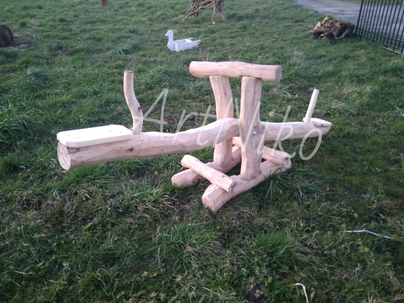 Drewniana huśtawka-ważka dla dzieci, wykonana z okrąglaków, umieszczona na trawniku. W tle widoczna dekoracja w kształcie kaczki.