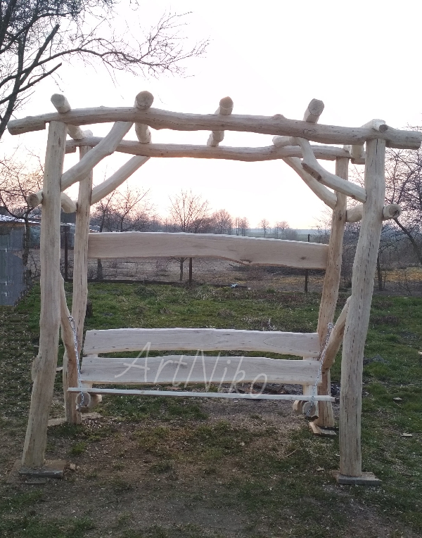 Drewniana huśtawka ogrodowa z surowych bali, zawieszona na łańcuchach, z widocznym logo 'ArtNiko' na siedzisku, ustawiona na trawniku w ogrodzie o zmierzchu.